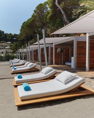 A row of modern cabanas offer seclusion and shade along a private private beach, with large white chaises and blue towels