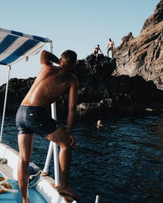 A tanned man, wet from a dip in the sea, leans on his boat's canopy bar as he chats to friends on the cliff and in the sea