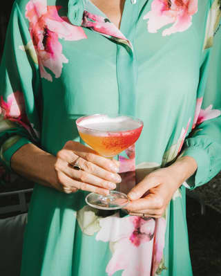 Two hands holding a peach cocktail in a coupe glass in front of a green, floral silk blouse