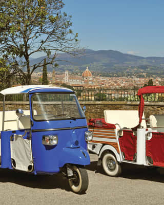 A red and white and blue and white three-wheel Ape stand waiting for passengers. Behind them are the red roofs of Florence