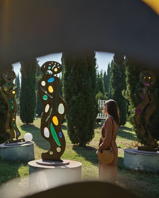 Lady in a tan dress strolling among art sculptures in a park lined with cypress trees
