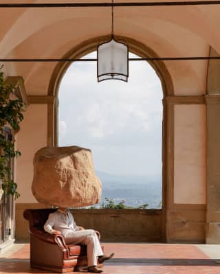 In an art installation a man sits a stone columned loggia, with Tuscan hills behind. In place of his head is a sandstone boulder