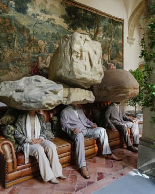 Two men and a woman sit on a sofa in front of a green fresco. Boulders take the place of their heads in this living sculpture