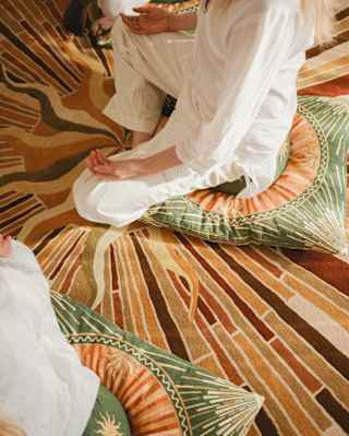 Two people dressed in white sit cross-legged on patterned cushions atop a brown and orange abstract rug. The photo is taken from above, showing only their lower bodies and hands in relaxed positions.