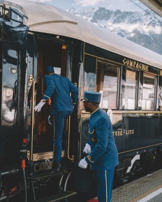 Two stewards in blue suits with gold trim, flat topped hats and white gloves board the train with the Alpine peaks behind