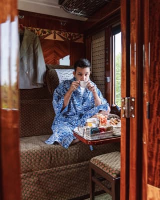 Man in a blue-patterned dressing gown sitting cross-legged and sipping tea in a vintage train cabin