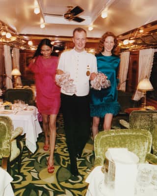 Smiling steward carrying crystal glasses and strolling through a train dining car with two ladies in dresses