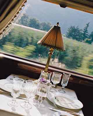 A green landscape rushes past a window framed by curtains next to an Etoile du Nord dining table, seen at an angle.