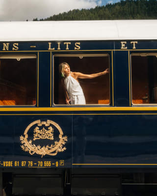 A blond woman in a white halter-neck dress stretches an arm across the train window as she peers out, seen from the platform.