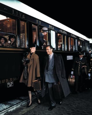 A couple dressed elegantly in coats and hat walk by the side of the carriage. Behind them gold-trimmed stewards carry bags
