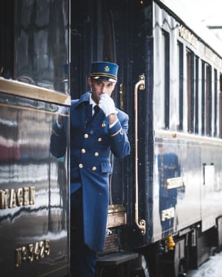 Train guard in a blue uniform blowing a whistle next to train carriages