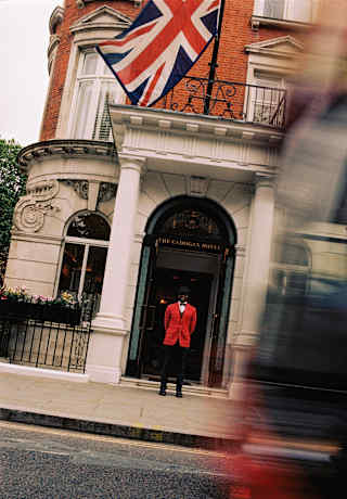 Tilted view across a busy road of a doorman in a red jacket with black trousers and bow tie outside the hotel entrance.