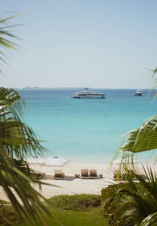 View through palms of the white sand beach where sun loungers face the sea and two yachts rest, suspended in the azure waters.