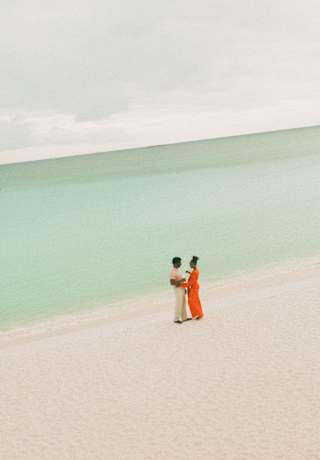 A man dances with a woman in bright orange playsuit which creates a vibrant splash on the white-sand beach, seen from afar.