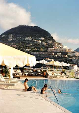 A boy sits with his feet in the pool as two girls play in the water in a northward view that captures a Ravello hillside.