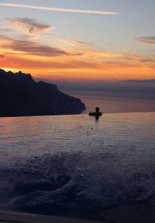 A swimmer rests at the edge of infinity pool watching the silhouetted coastline melt into the purple haze beneath a sunset.