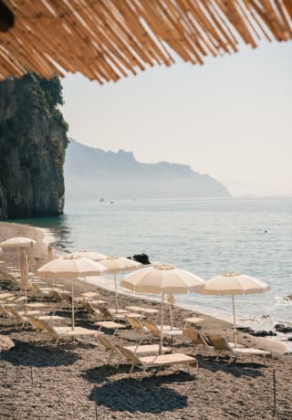 Two pristine rows of chaise longues lie facing the sea on a private pebble beach, each pair shaded by a white parasol