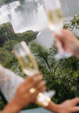 Two guests raise champagne flutes, seen in close-up in soft-focus foreground, with a dramatic Iguassu Falls backdrop.