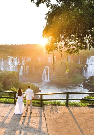 A man in white shirt and woman in long white dress hold hands gazing at the cascades of Iguassu Falls