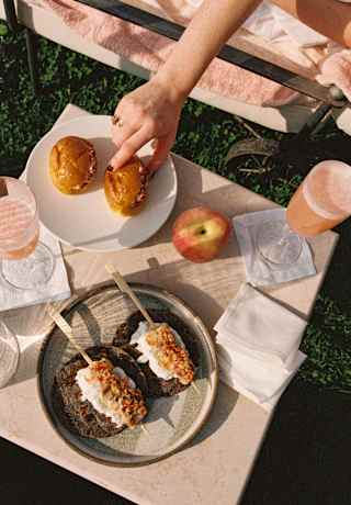 A woman's hand reaches for one of two paninis at a table with Peach Bellinis and savoury skewers on bread, seen from above.