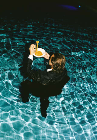 A tuxedo-dressed man stands partially submerged in a swimming pool, a fork of spaghetti held aloft above his plate of pasta