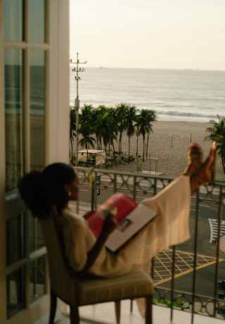 Seen in soft-focus, a woman reads a magazine on a chair with her feet up on the railings of her beach-view balcony.