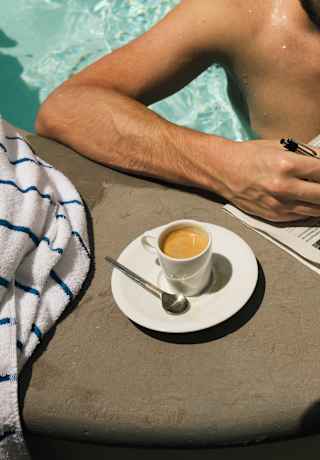 A man standing in the pool completes a crossword puzzle at the poolside, alongside a towel and a cup of coffee with a saucer.