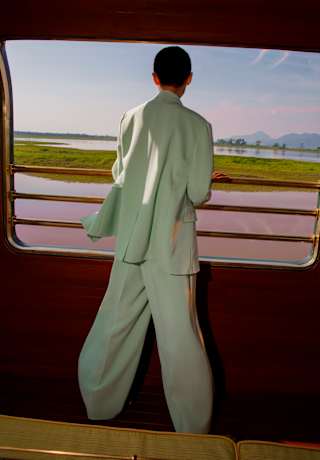 A woman in a pale mint trouser suit looks out from an open Observation Car window at wetlands in low sun, seen from behind.