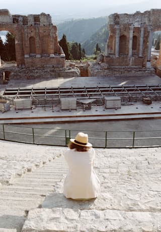 A women in a white jacket and panama hat sits on the stone steps of the Greek theatre as technicians build a stage below