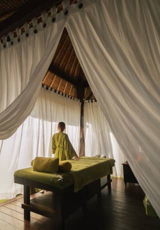 A woman in an olive-green gown stands by a treatment bed at the Beach Spa, seen from behind, through open white drapes.>
