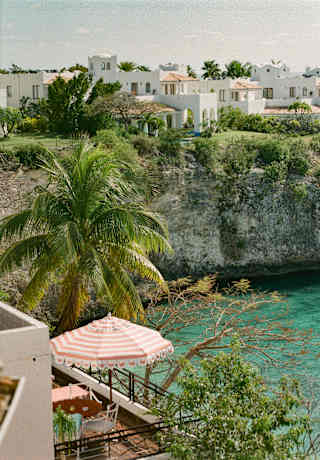 View over a terrace with a red and white-striped parasol to aquamarine waters, overlooked by La Samanna on the cliff above.
