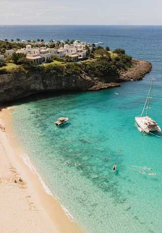 Four boats float in Baie Longue's sapphire waters, sheltered by a headland dotted with resort residences, in an aerial view.