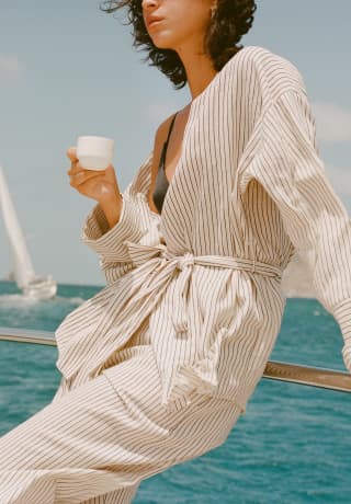 A woman enjoys an onboard espresso during a day at sea, as a keeling sailboat behind glides through the aquamarine seascape.