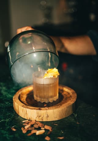 A bartender lifts a glass, smoke-filled cloche from a wood base, revealing a Smoked Old Fashioned cocktail at Bar Bambuco.