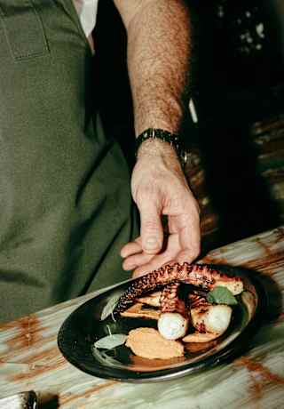 A person in an apron is holding a black plate with grilled octopus tentacles, scallops, and garnish on a rustic table.