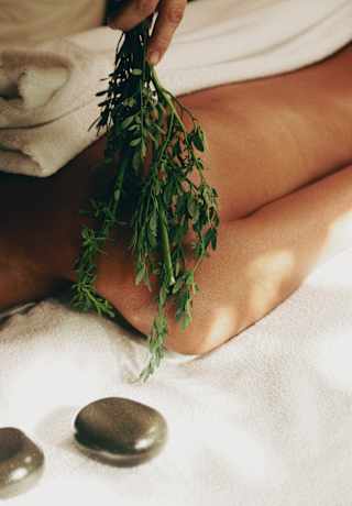Close-up of a guest's shoulder as a spa therapist strokes the skin with herbs, in a  detail capturing two massage stones.>