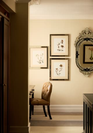 Above a desk with leather chair, three botanical prints hang on a wall alongside an oval mirror, glimpsed through a doorway.