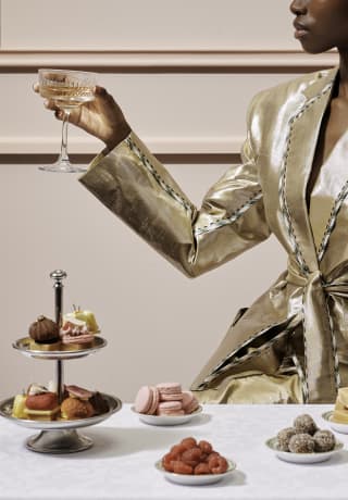 A woman in a gold suit holds a champagne glass at a high-tea table with a silver cake stand and plates of maracons and fruit.