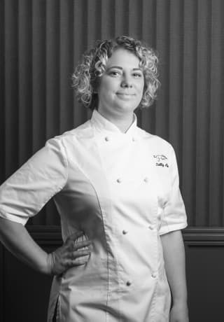 A black and white image of acclaimed British chef, Sally Abé, as she poses for the camera in chef's whites with her hand on hip