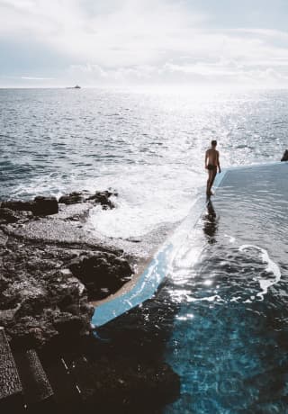 A man in a bathing suit stands on the edge of the saltwater pool, looking out over the sparkling Atlantic ocean.