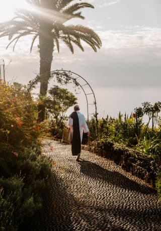 A guest in a white robe with a towel on one shoulder casts a shadow behind him as he walks on a garden path with sea views.>