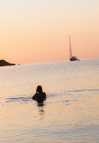 A swimmer stands in chest-high water tinged silver and peach by the sunset which glows on the horizon, silhouetting a boat.