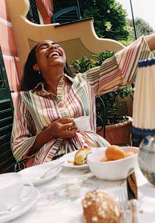 A woman in a candy-stripe top holds a coffee cup and tips her head back to laugh in an angled view over a breakfast table.