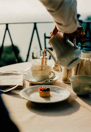 A waiter pours tea from a pot at a table with cutlery, napkins and a fruit tartlet on a plate, in a breakfast detail.