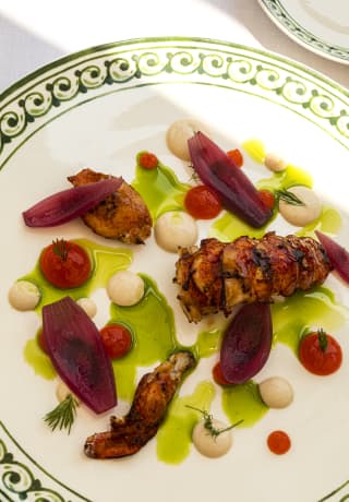 Grilled lobster tail and claws, served with red onion and a green, herby oil on a white plate with a green patterned rim.
