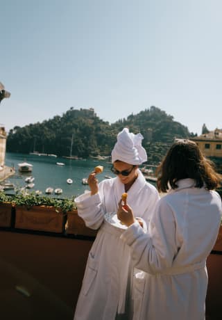 Two women in bathrobes eating and laughing on a balcony, Portofino harbour in the background