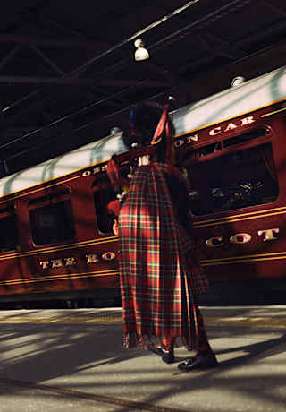 Half-hidden in shadow, a bagpipe player in traditional kilt, plays on a platform in front of the stationary train.