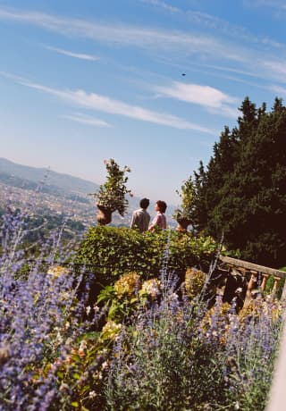 Angled image of two guests standing on the stone terrace in a garden bursting with lavender and foliage overlooking Florence.