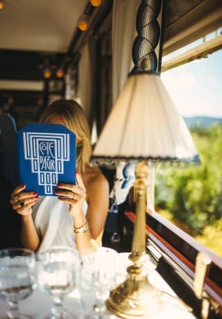 Seen across a table with a lamp and three glasses, a woman holds a blue Cote d'Azur Dining Car menu in front of her face.