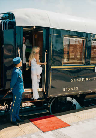 A Pullman Porter in blue watches as a female guest in a white trouser suit with long blond hair mounts the carriage steps.